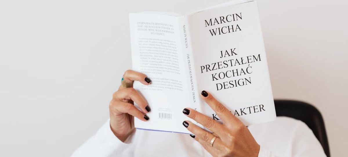 Anonymous female in white shirt with gold chain on neck reading interesting book covering face while sitting on black chair
