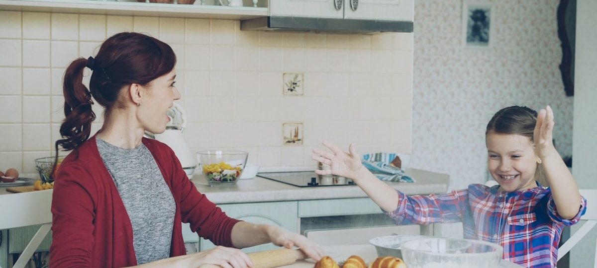 Young mother and cute daughter cooking together talking in the kitchen on weekend. Little funny girl clapping hands with flour and laughing. Family, food, home and people concept
